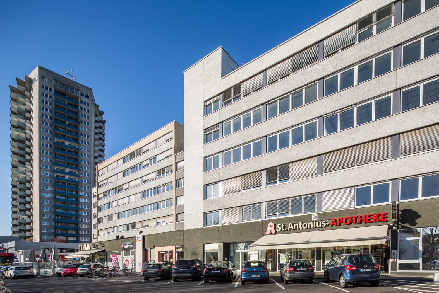 Langgestrecktes mehrgeschossiges Gebäude mit Apotheke und Ladenzeile im Erdgeschoss, davor ein Parkplatz, links ein Wohnhochhaus.