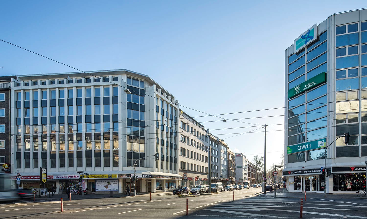 Innerstädtische Kreuzung mit einem hellen Geschäftshaus links und einem verglasten Eckgebäude rechts, dazwischen Straßenbahnoberleitungen.