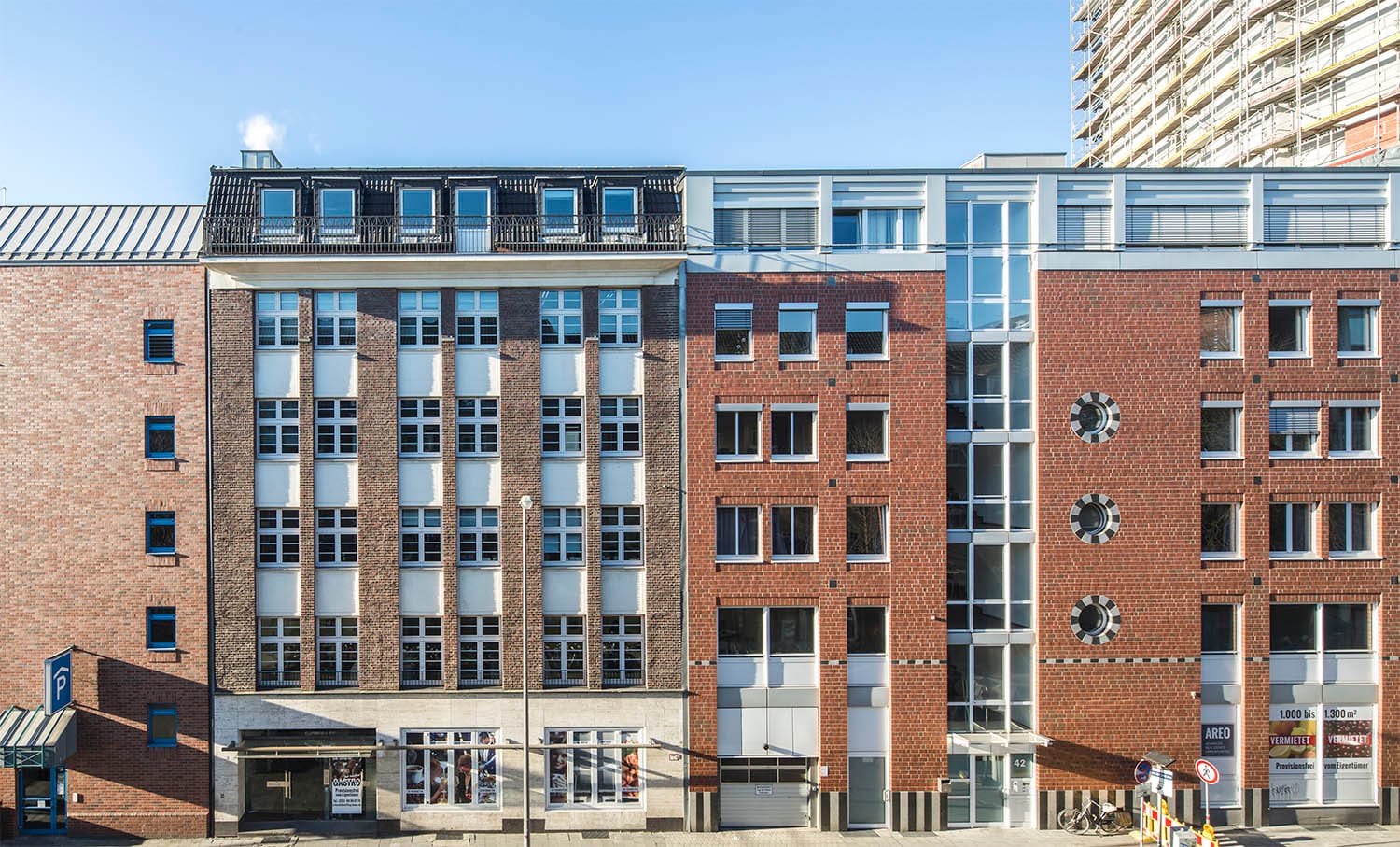 Straßenzeile aus Backsteinhäusern mit hellen Fensterachsen und dunklem Staffelgeschoss, im Hintergrund ein eingerüstetes Hochhaus.