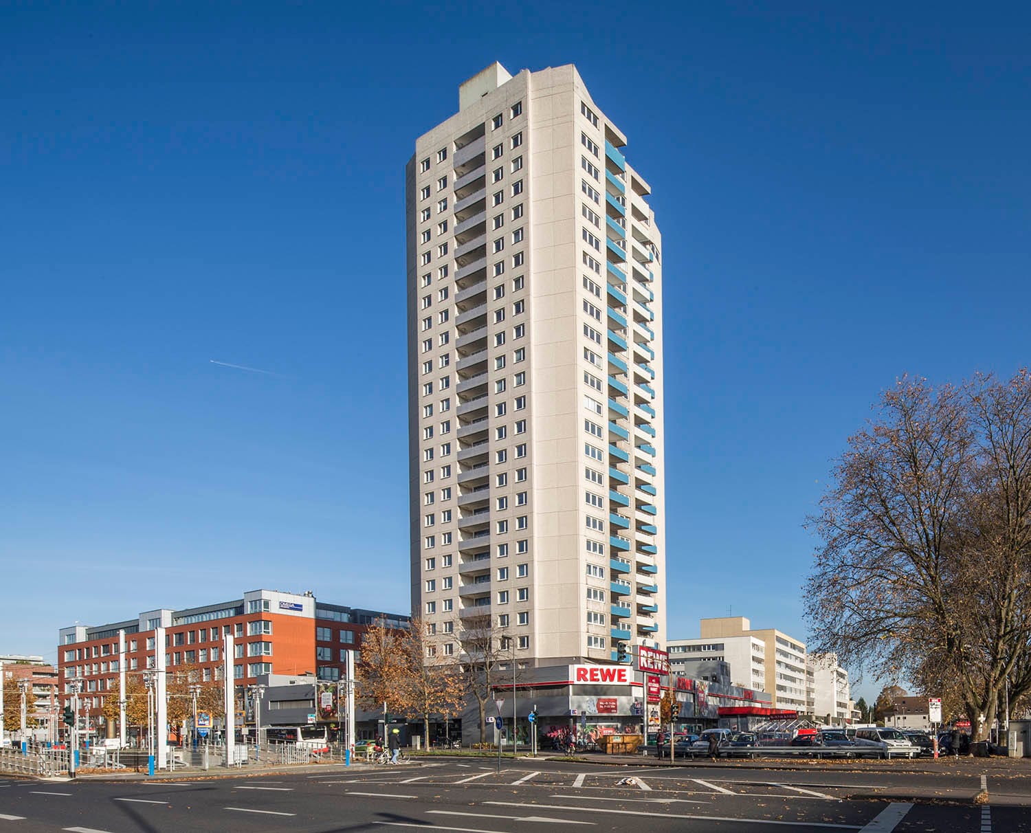 Schlankes Wohnhochhaus mit vorspringenden Balkonen über einer eingeschossigen Ladenzeile, davor eine breite Straßenkreuzung.